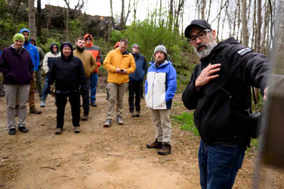 A man explaining trail signage to a big group standing in a circle in the woods