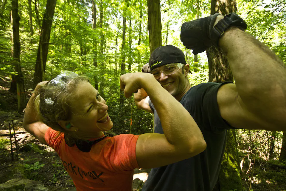 A man and woman flexing their muscles and smiling while in the woods