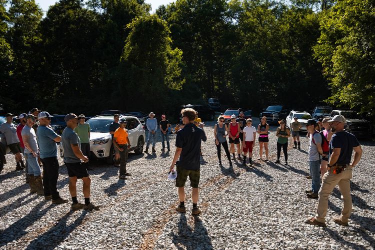 A man speaks to a group in a gravel parking lot outdoors